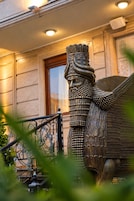 Une statue en bronze représentant une figure ailée, coiffée d’un headdress finement travaillé, est placée devant un bâtiment doté d’une porte en bois et d’une rambarde en métal.
