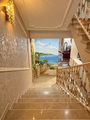 A grand staircase with a view of a painted seascape, leading to a room with a red sofa and a chandelier. - Lamassu Luxury Suites Batumi (Batumi)