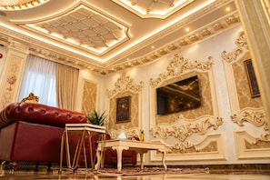 Una habitación lujosa con un sofá rojo, un televisor con marco dorado, decoraciones murales ornamentales y un candelabro.