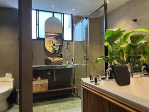 A modern bathroom with a glass-enclosed shower, a round mirror, a sink with a stone basin, and a large potted plant. - Luxury Sense I (Medellín)