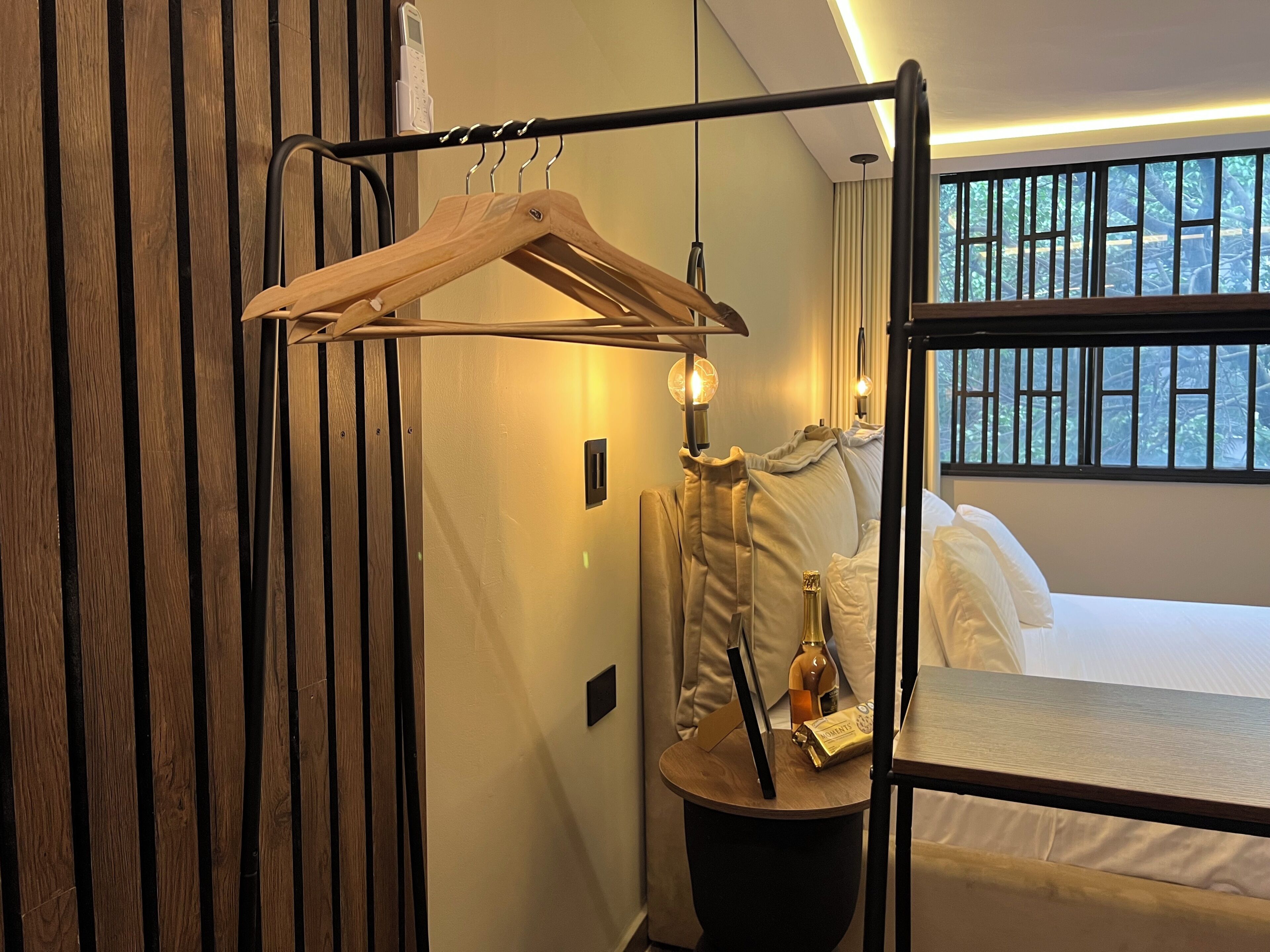 Ein Hotelzimmer mit Etagenbett, einem Holzgalgen an einem Metallgestell, einem Nachttisch mit Flasche und Buch sowie einem Fenster mit Blick auf Bäume.
