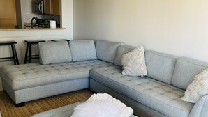 A modern living room with a grey sectional sofa, a white pouf, and a kitchen area in the background.