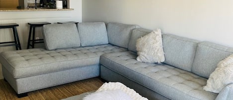 A modern living room with a grey sectional sofa, a white pouf, and a kitchen area in the background.