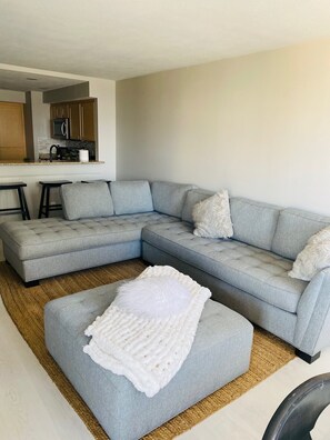 A modern living room with a grey sectional sofa, a white pouf, and a kitchen area in the background.