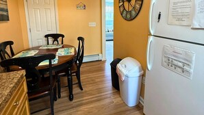 A kitchen with a dining table, chairs, a refrigerator, a trash bin, and a wall clock.