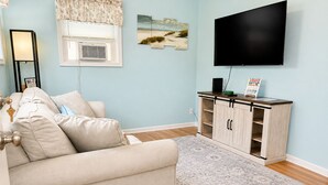 A living room with a beige sofa, a flat-screen TV mounted on the wall, a wooden cabinet with a tray, and a patterned rug.
