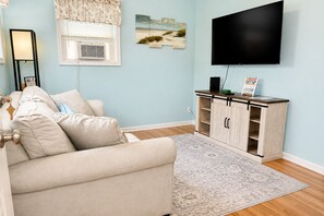 A living room with a beige sofa, a flat-screen TV mounted on the wall, a wooden cabinet with a tray, and a patterned rug.