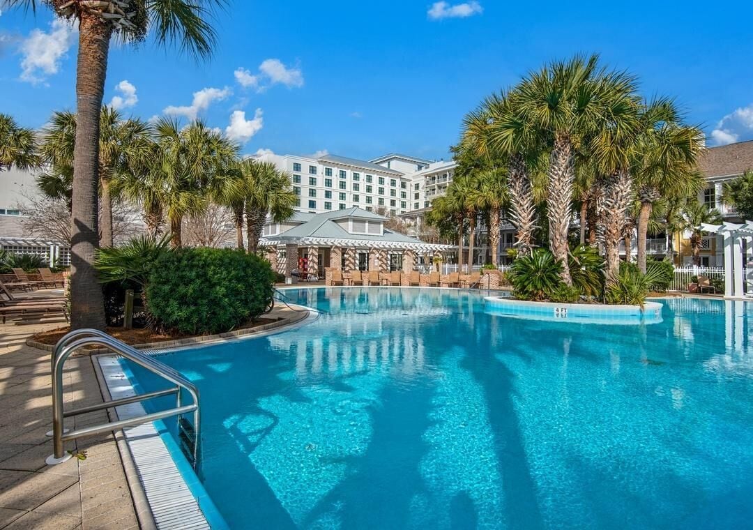 A hotel pool area with a clear blue pool, palm trees, and a building with a white facade in the background.