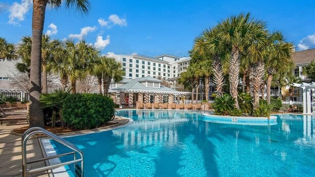 Área de piscina de hotel com uma piscina azul cristalina, palmeiras e um edifício com fachada branca ao fundo.