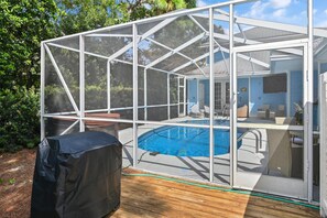 A covered outdoor swimming pool with a wooden deck, surrounded by greenery. - Blue Chip Escape (Santa Rosa Beach)