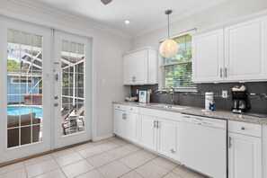 A modern kitchen with white cabinetry, a granite countertop, and a view of a swimming pool through glass doors. - Blue Chip Escape (Santa Rosa Beach)
