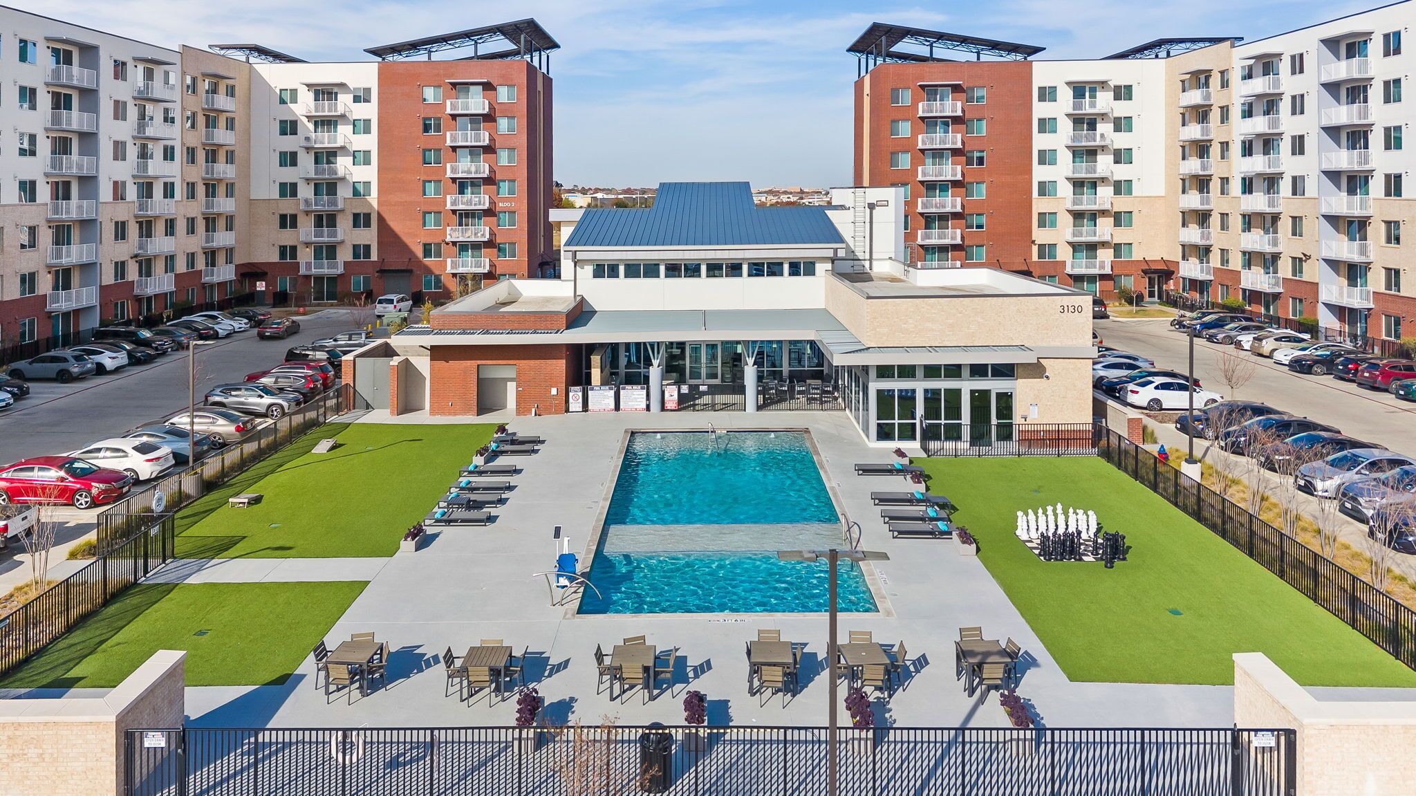 A modern apartment complex with a central swimming pool, outdoor seating, and a parking area.