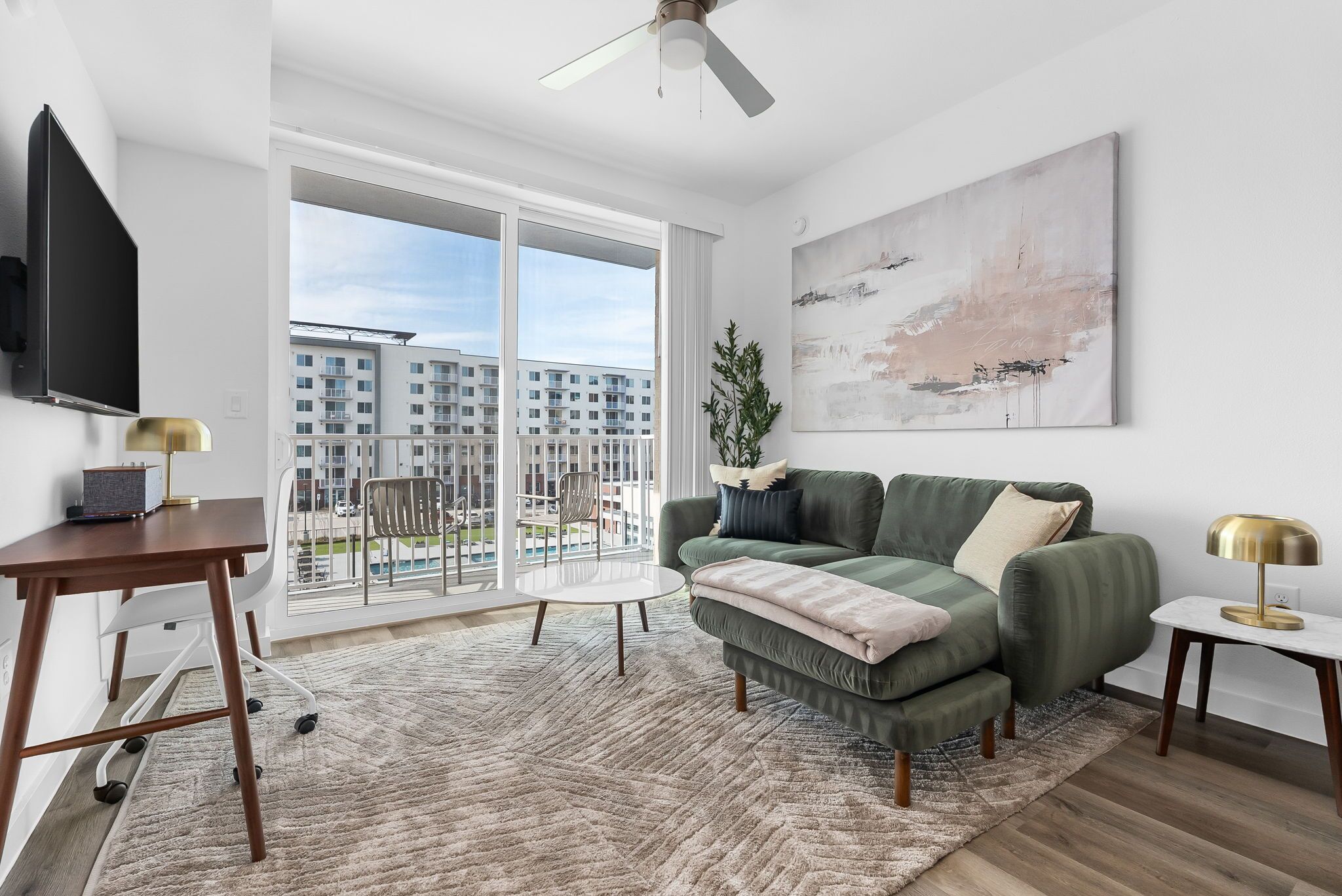 A modern living room with a green sofa, a coffee table, a TV, and a balcony with a view of apartment buildings.