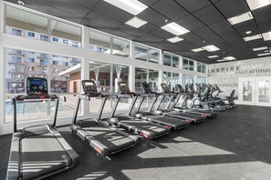 A gym with treadmills aligned in rows, large windows, and a view of buildings outside.