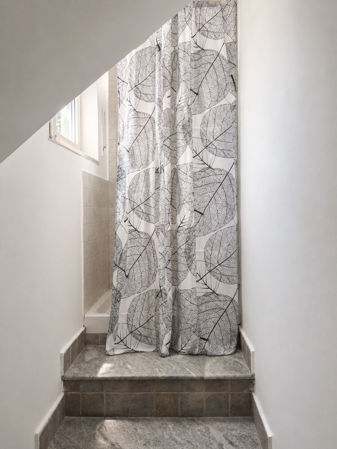 Un bagno con tenda da doccia decorata con un motivo a foglia, zona doccia piastrellata e una finestra sopra la doccia.
