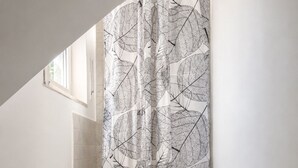 Ein Badezimmer mit Duschvorhang mit Blättermotiv, einer gefliesten Duschkabine und einem Fenster über der Dusche.