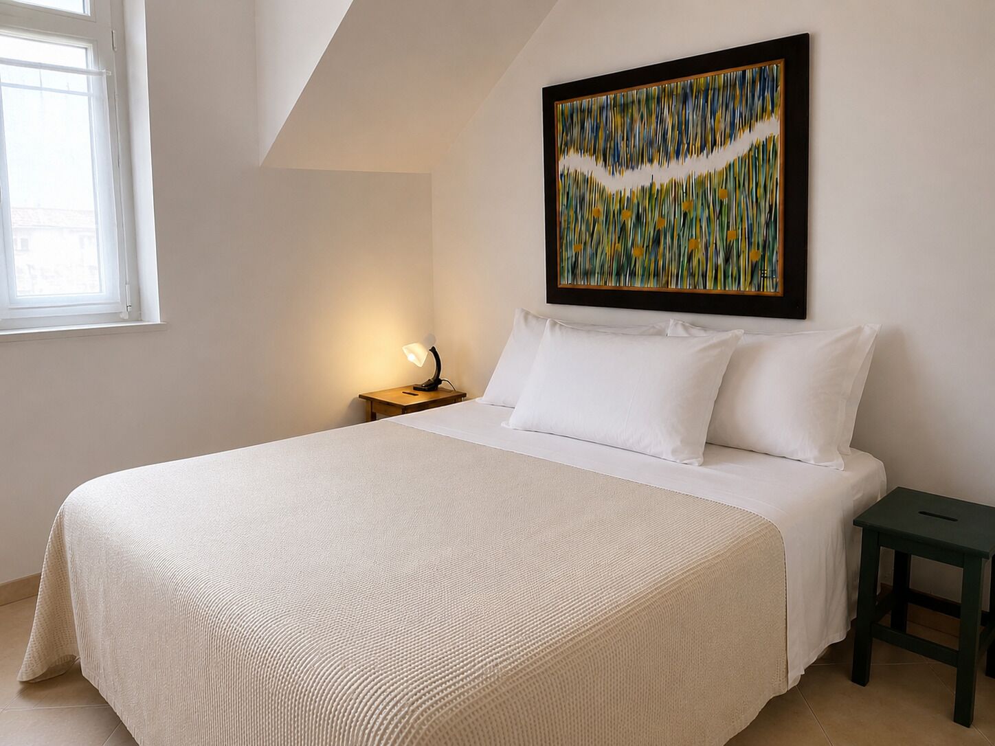 Una camera da letto con un letto grande, un comodino con una lampada, un quadro astratto incorniciato appeso al muro e una finestra che lascia entrare la luce del giorno.