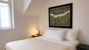 Ein Schlafzimmer mit einem großen Bett, einem Nachttisch mit Lampe, einem gerahmten abstrakten Gemälde an der Wand und einem Fenster mit Tageslicht.