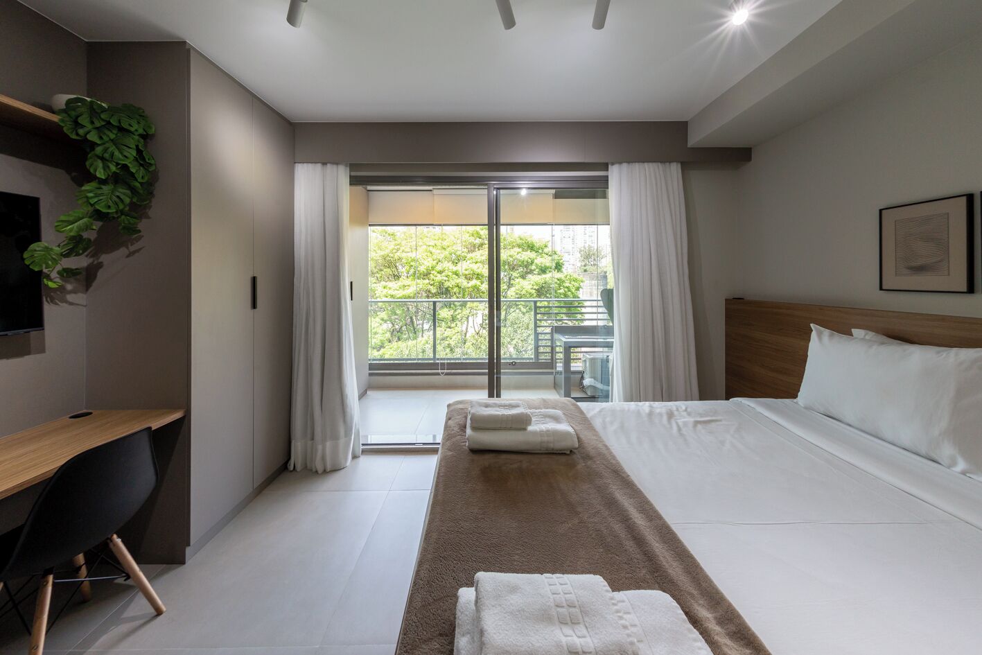 A hotel room with a bed, a desk, a chair, a TV, a window with curtains, and a balcony with greenery.
