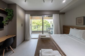 A hotel room with a bed, a desk, a chair, a TV, a window with curtains, and a balcony with greenery. - Caraibas by Bhomy (São Paulo)