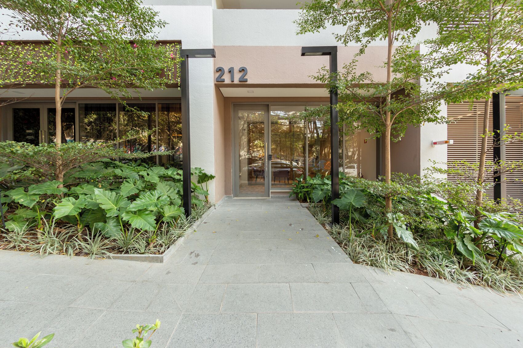 A building entrance with a number '212' above the door, surrounded by greenery and a paved walkway.