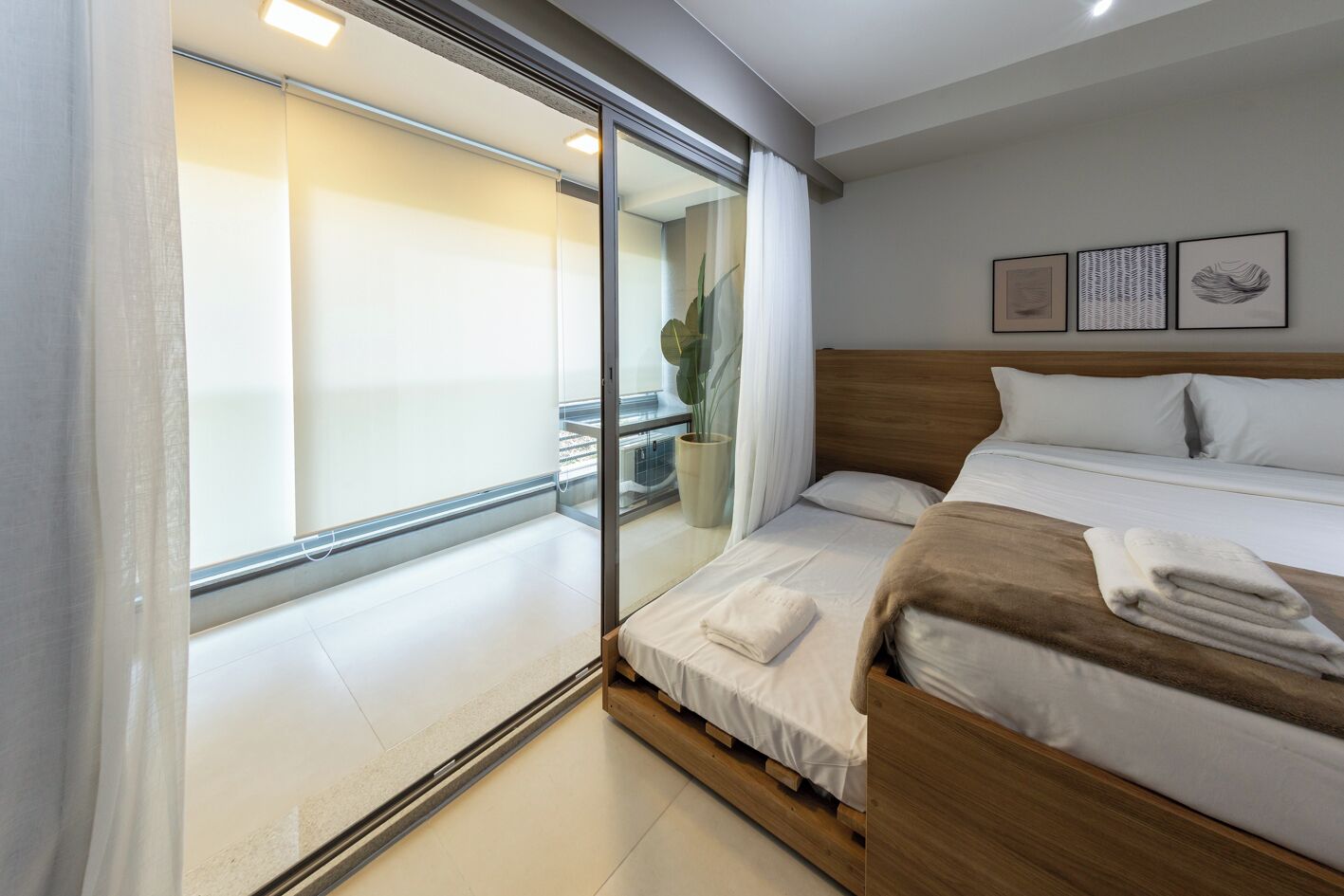 A hotel room with a bed, a bedside table, a potted plant, and a view of the balcony.