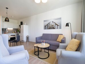 Ein modernes Wohnzimmer mit einer Couch, zwei Sesseln, einem kleinen Couchtisch und einer Küche im Hintergrund.
