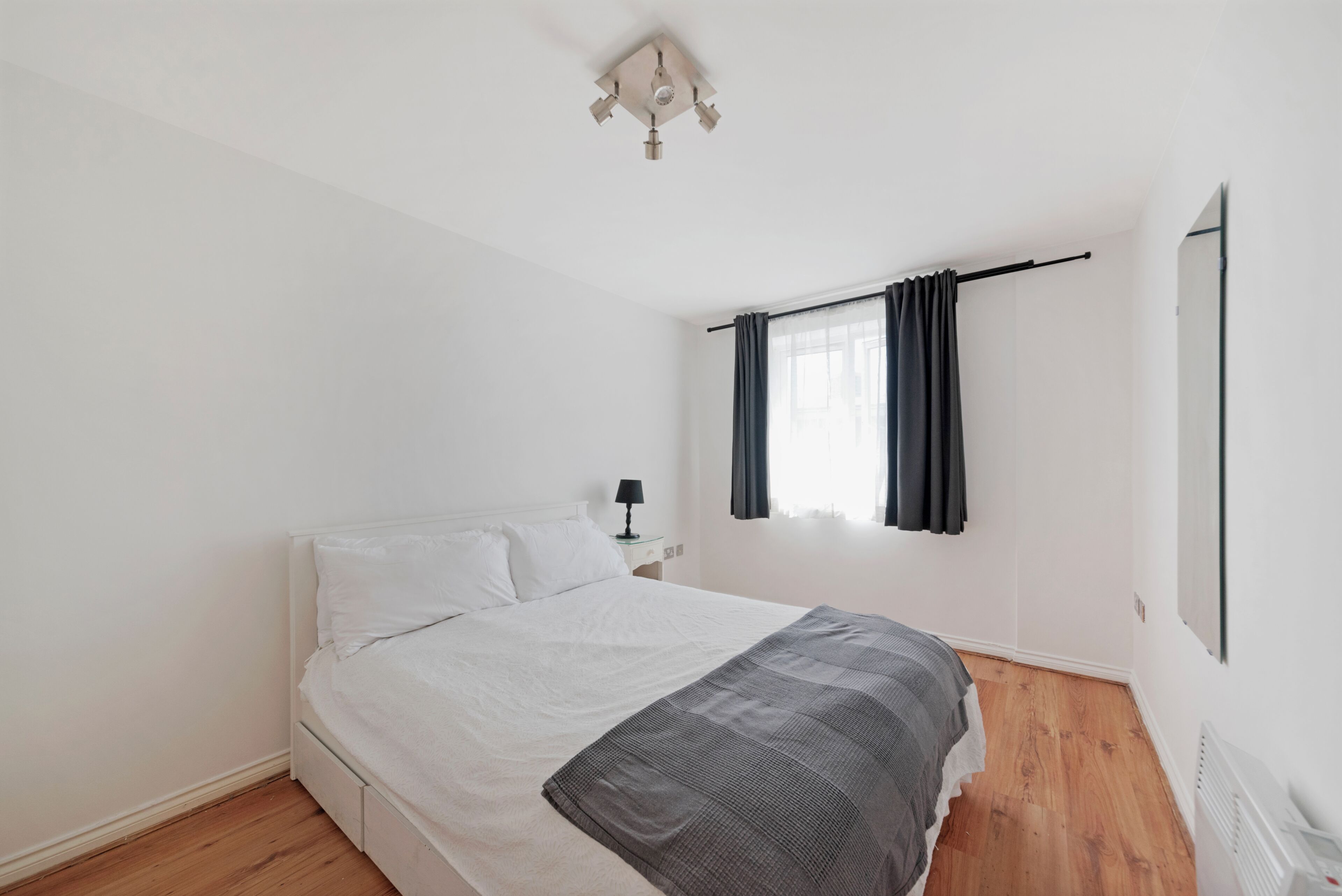 Une chambre à coucher avec un lit, une table de chevet, une lampe, une fenêtre avec des rideaux et un radiateur.