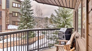 A balcony with a grill, a chair, and a view of a snow-covered landscape with trees. - Walk to Lifts & Main St | Hot Tub (Breckenridge)