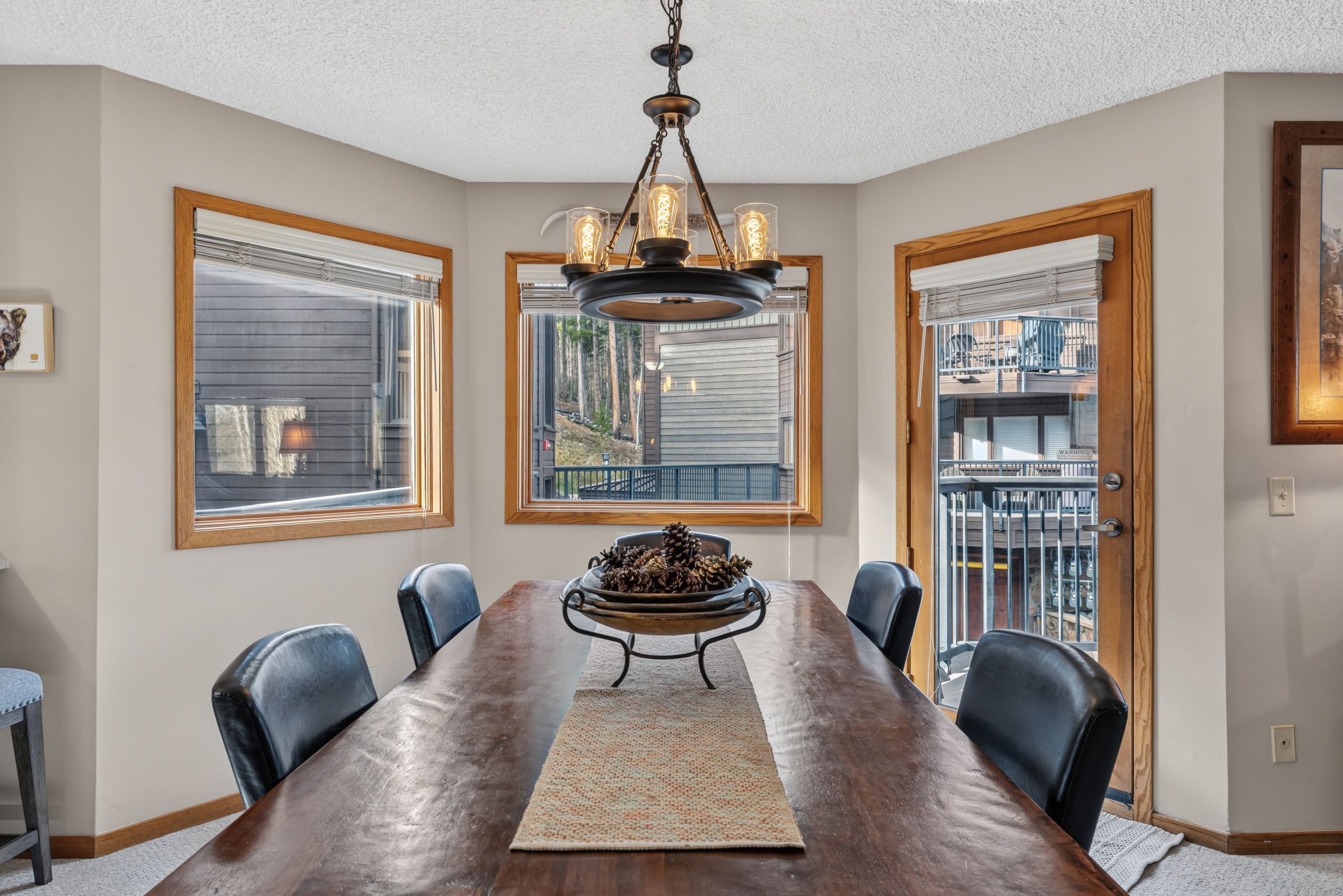 A dining room with a wooden table, chairs, a chandelier, and large windows with blinds.
