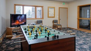 A room with a foosball table, a television displaying a sports game, and a window with a view of trees. - Walk to Lifts & Main St | Hot Tub (Breckenridge)