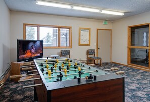 A room with a foosball table, a television displaying a sports game, and a window with a view of trees. - Walk to Lifts & Main St | Hot Tub (Breckenridge)