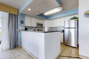 A modern kitchen with white cabinets, a stainless steel refrigerator, and a central island with a microwave. - Carolinian Beach Resort 0933 (Myrtle Beach)
