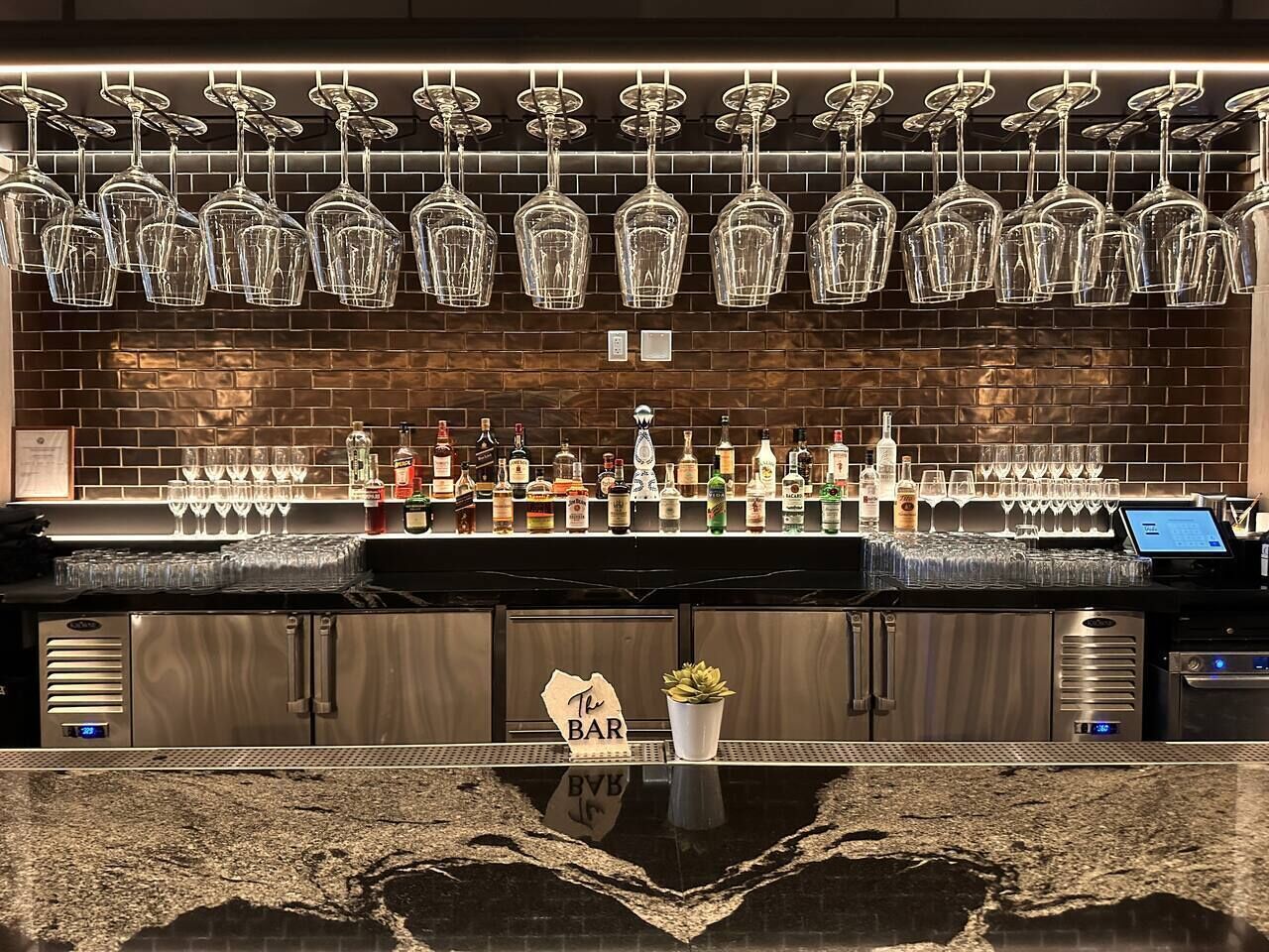 A bar area with a brick wall, hanging wine glasses, and a variety of liquor bottles.