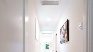 Um corredor comprido e estreito, com paredes brancas e chão de madeira clara. Há quadros nas paredes, um respiradouro no teto e uma porta no final do corredor.