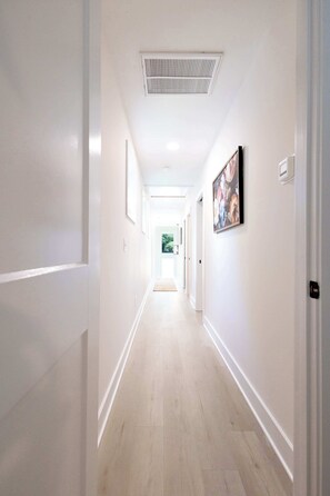 A long, narrow hallway with white walls and a light wooden floor. There are framed pictures on the walls, a vent on the ceiling, and a door at the end of the hallway. - 8 Min to Duke + Downtown | Back Deck (Durham)