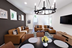 A modern living room with a dark brown wall, a large flat-screen TV, a ceiling fan, and a dining area with a round table. - 8 Min to Duke + Downtown | Back Deck (Durham)