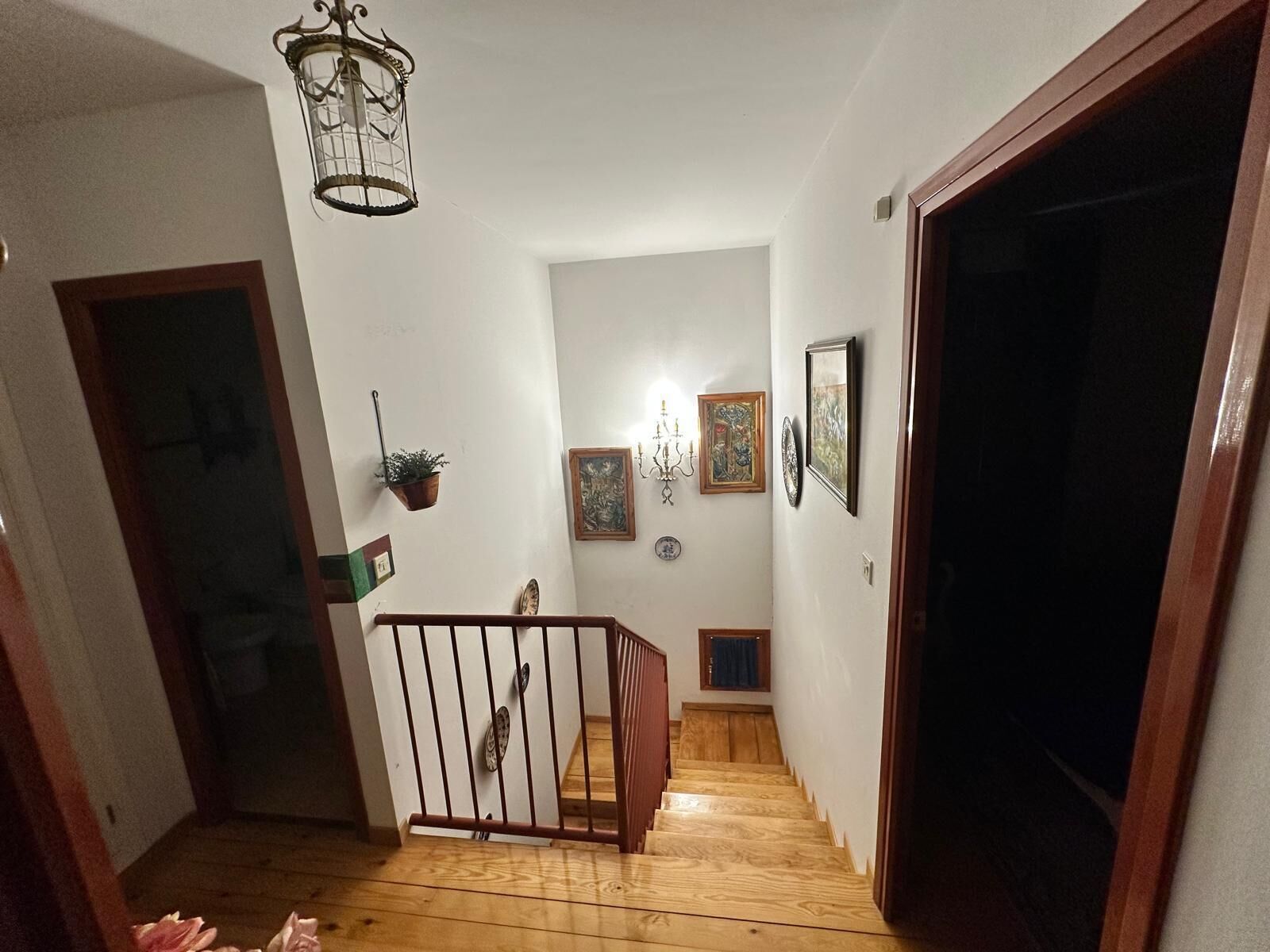 A wooden staircase leading to a landing with a chandelier, framed artwork, and a potted plant.