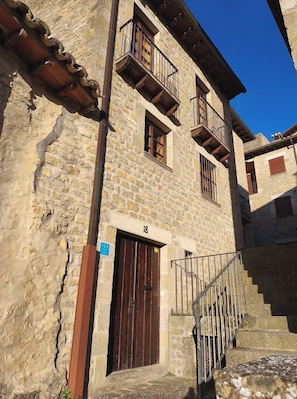 A stone building with a wooden door, a balcony, and a staircase leading to an upper level.