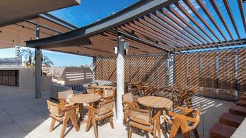 An outdoor seating area with wooden tables and chairs, a pergola structure, and a view of the sky.