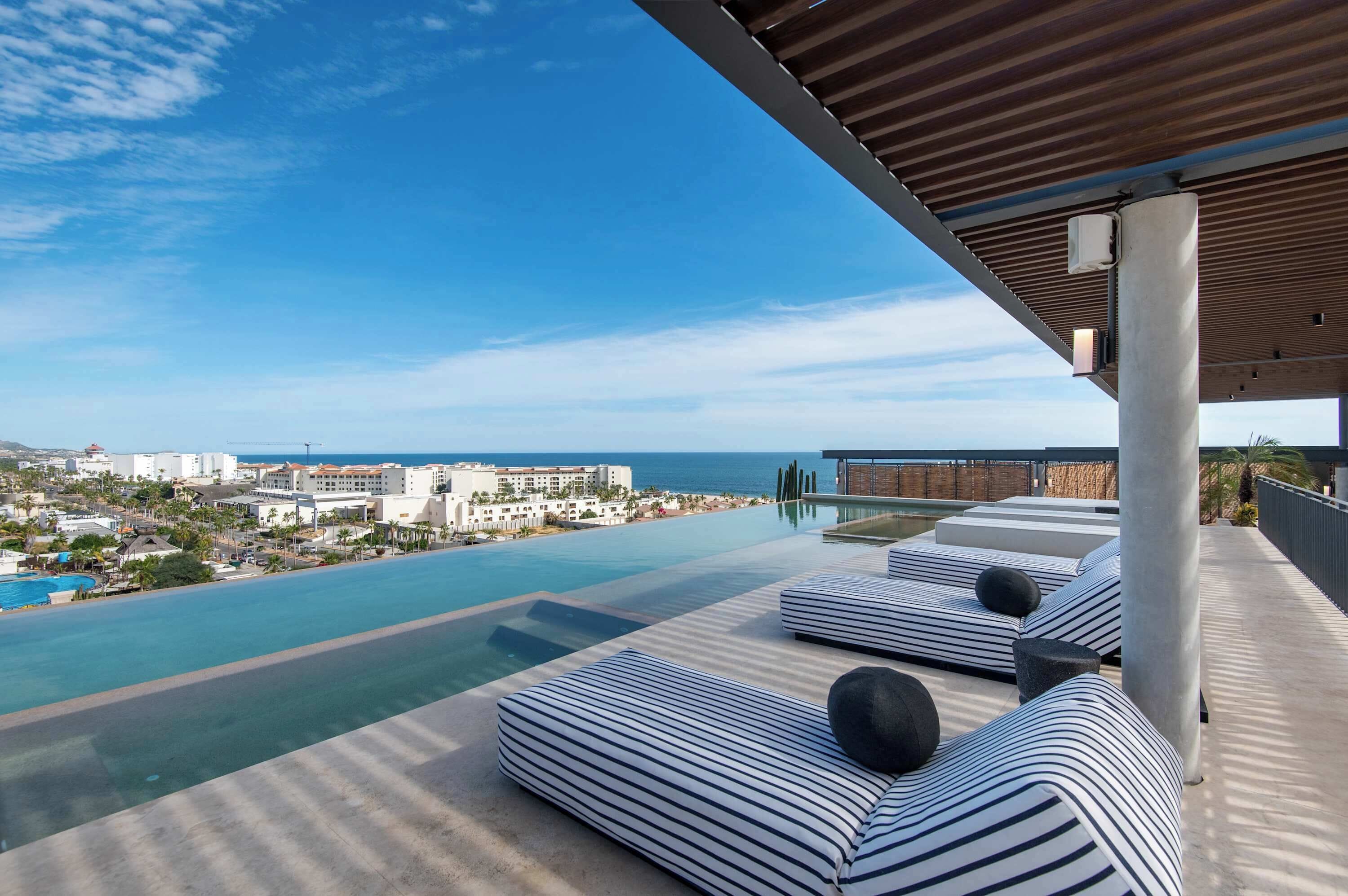 Ein Rooftop-Infinity-Pool mit gestreiften Liegestühlen und Blick auf Stadt und Meer.