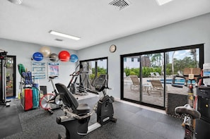 A gym with exercise equipment, a wall clock, and a view of a pool area through large windows. - Sunset at Banyan Bay|Gulf-Front Condo on Longboat Key Beach+Pool+Tennis |Sleep 8 (Longboat Key)