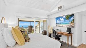 Een slaapkamer met een bed, een bureau, een computer en een groot raam met uitzicht op het strand.