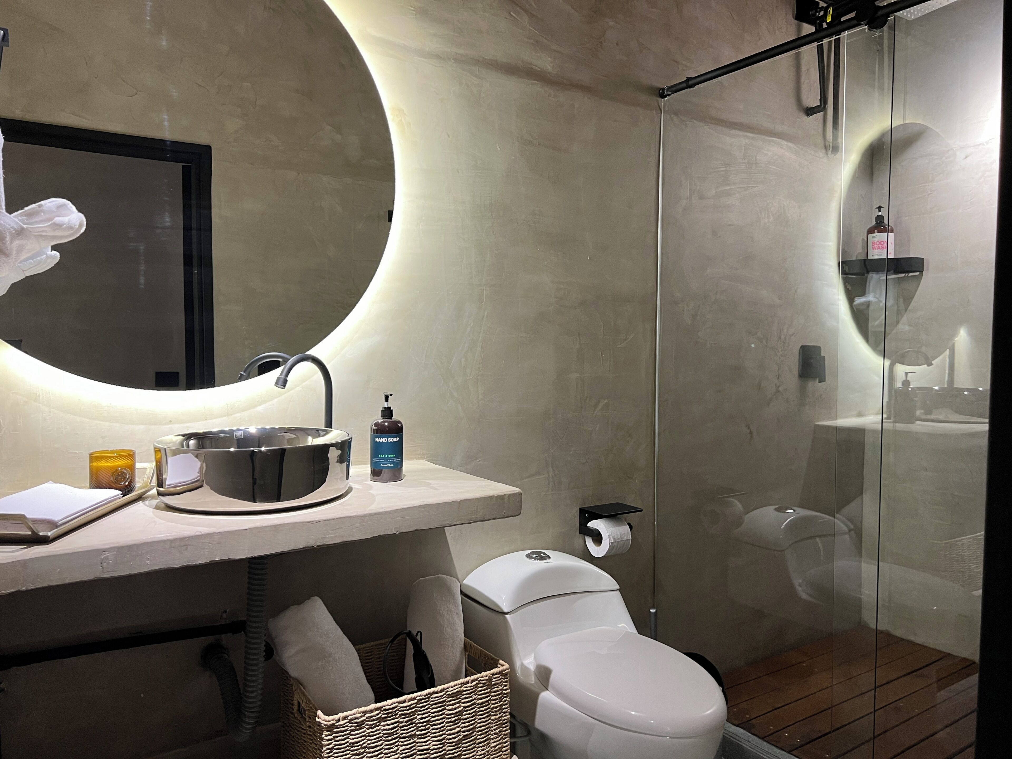 Ein modernes Badezimmer mit rundem Spiegel, Waschbecken mit Seifenspender, Toilette und einer Duschkabine aus Glas.