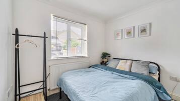 A bedroom with a bed, a radiator, a window with blinds, a coat rack, and a wall with framed pictures.