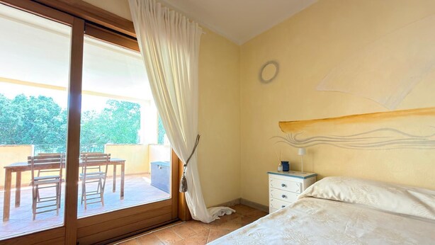 A bedroom with a bed, a nightstand, a window with curtains, and a view of an outdoor area. - Ginestre Smeraldo by Casa Check (Porto Rotondo)