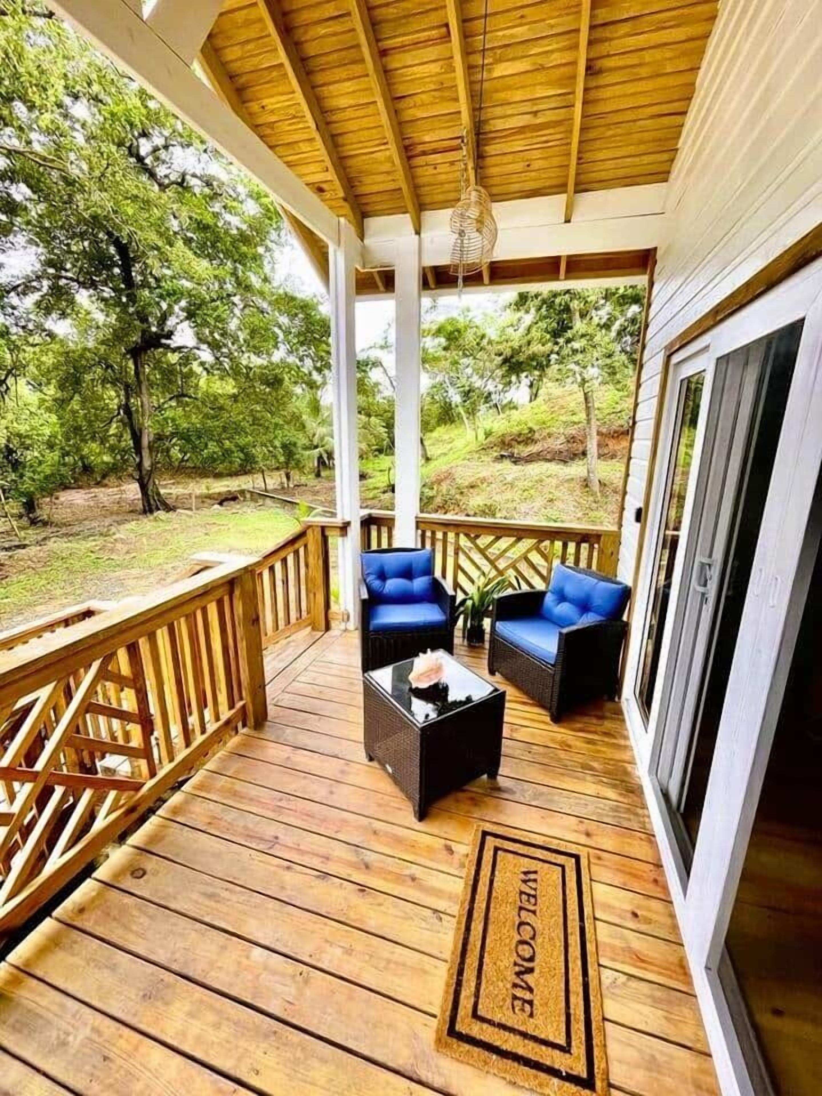 A wooden deck with two blue chairs and a coffee table, a welcome mat, and a view of trees.