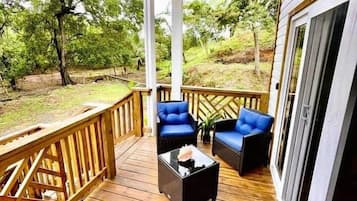 A wooden deck with two blue chairs and a coffee table, a welcome mat, and a view of trees.