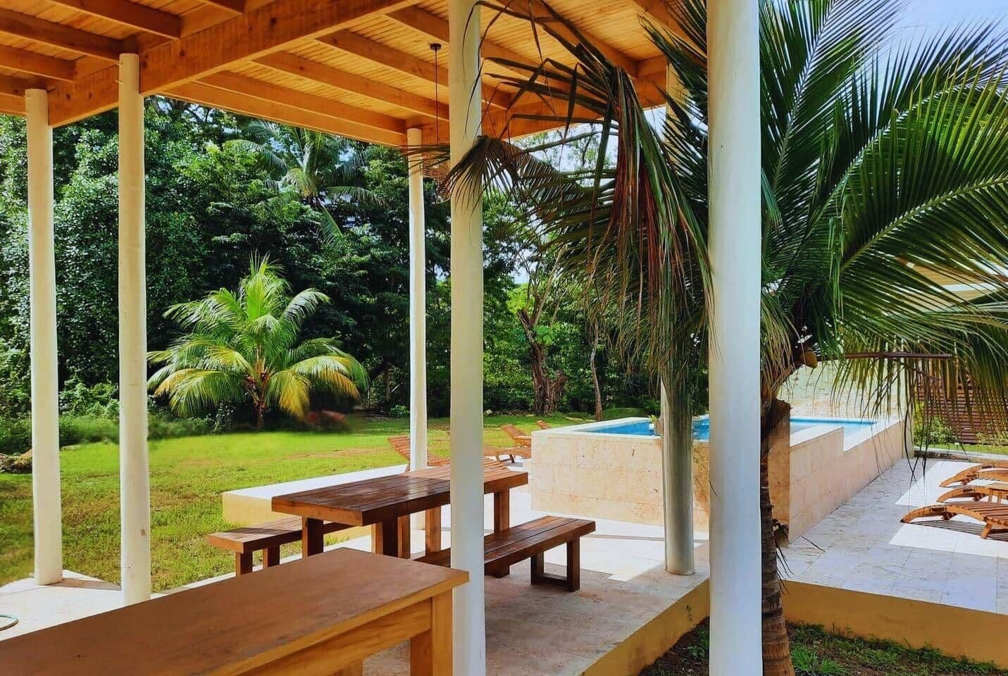 Een terras buiten met houten tafels en banken, omgeven door weelderig groen en een zwembad.