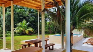 An outdoor patio with wooden tables and benches, surrounded by lush greenery and a swimming pool. - Conch Casitas - King or 2 Singles, AC, Shared Pool (Roatán)
